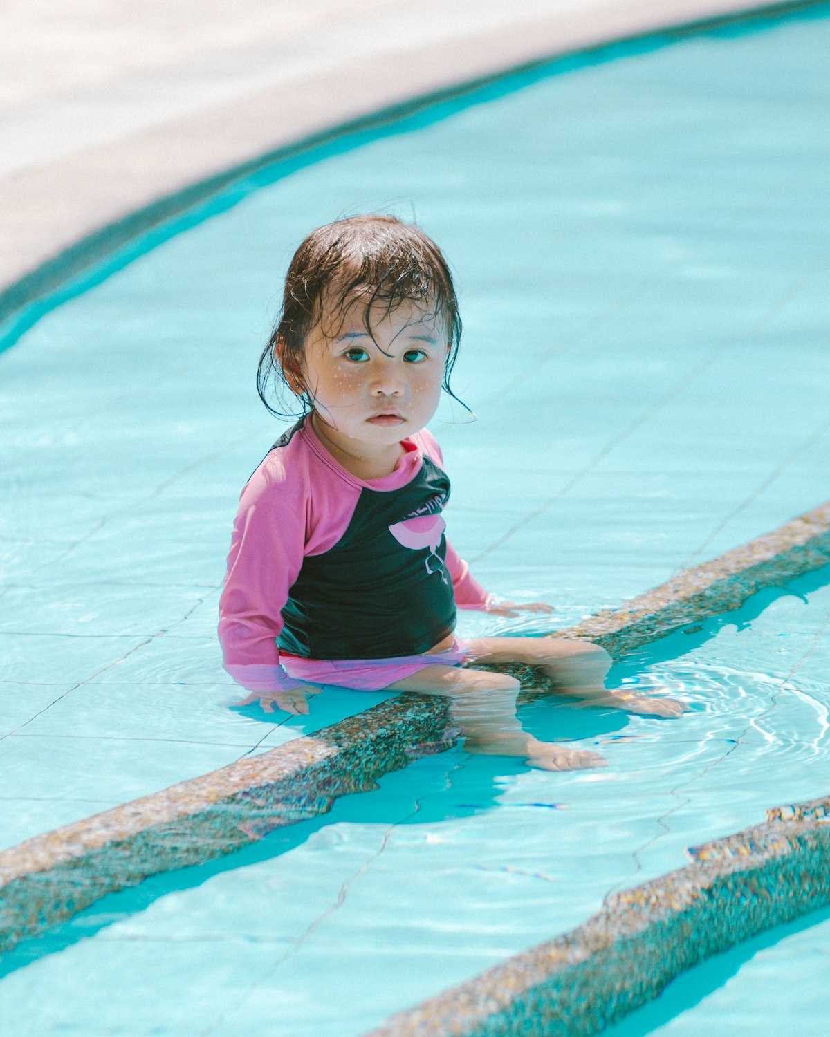 A toddler in a pink swimsuit sitting on the pool step